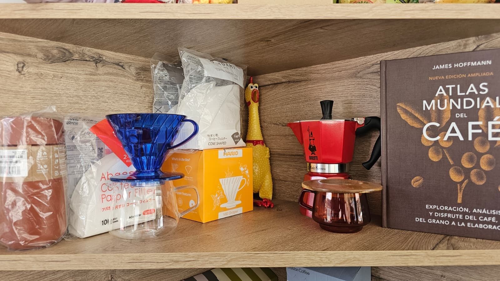 Estantería con un V60 azul, filtros de papel, una cafetera italiana roja sobre una jarra y un libro sobre café